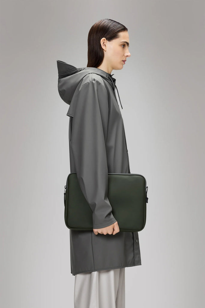 Person wearing a gray raincoat holding a green bag on a plain background. Rains Trail Laptop Case in Green.