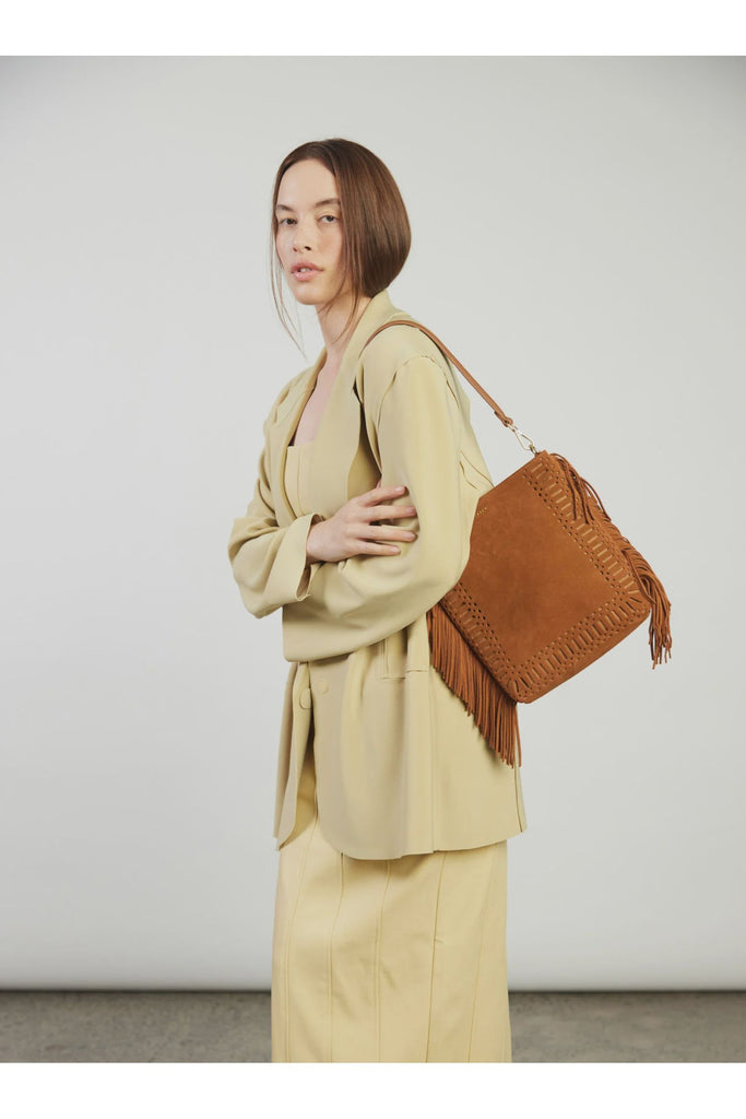 Woman in a beige suit holding a brown suede fringed handbag against a plain background. Saben Claudette Fringed Tassel Crossbody Bag in Tan Suede.