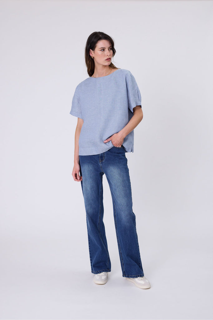 Woman wearing a light blue top and blue jeans on a white background. Marlow Sable Linen Top Blue Smoke.