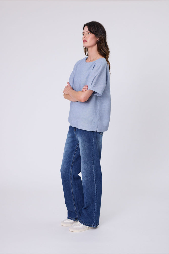 Woman wearing a light blue top and blue jeans on a white background. Marlow Sable Linen Top Blue Smoke.