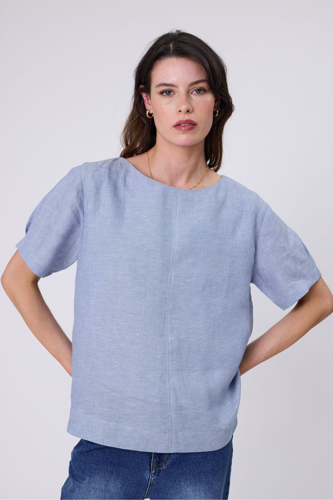 Woman wearing a light blue top and blue jeans on a white background. Marlow Sable Linen Top Blue Smoke.