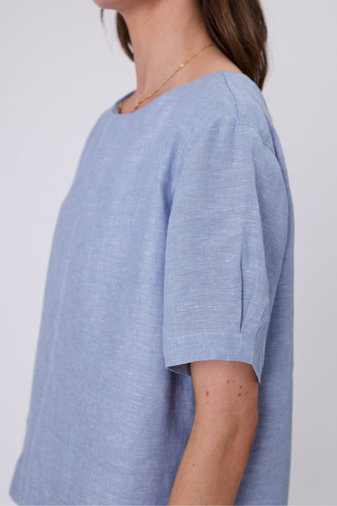 Woman wearing a light blue top and blue jeans on a white background. Marlow Sable Linen Top Blue Smoke.