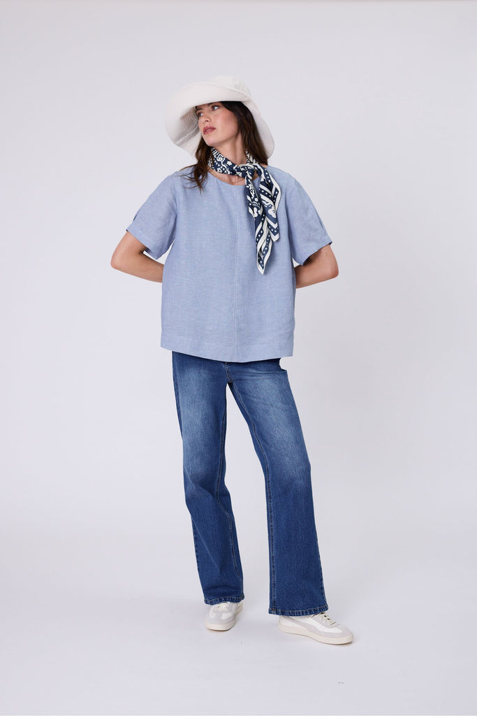 Woman wearing a light blue top and blue jeans on a white background. Marlow Sable Linen Top Blue Smoke.