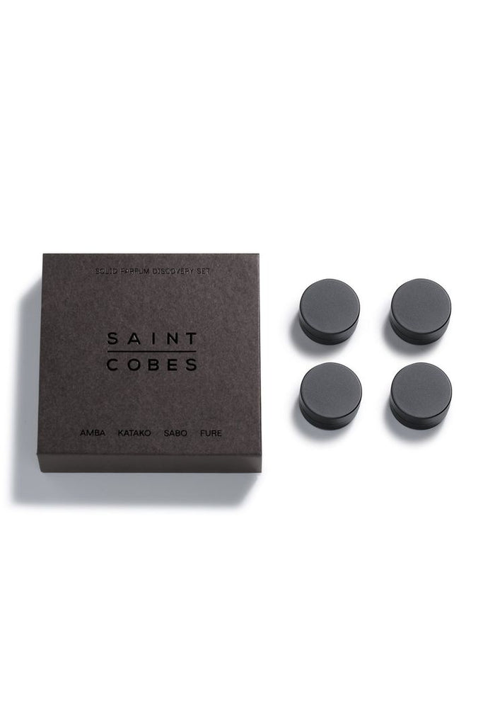 Saint Cobes Discovery Set. Set of four round black pottles containing solid fragrances sitting next to the black Saint Cobes box that the pottles are presented in on a white background