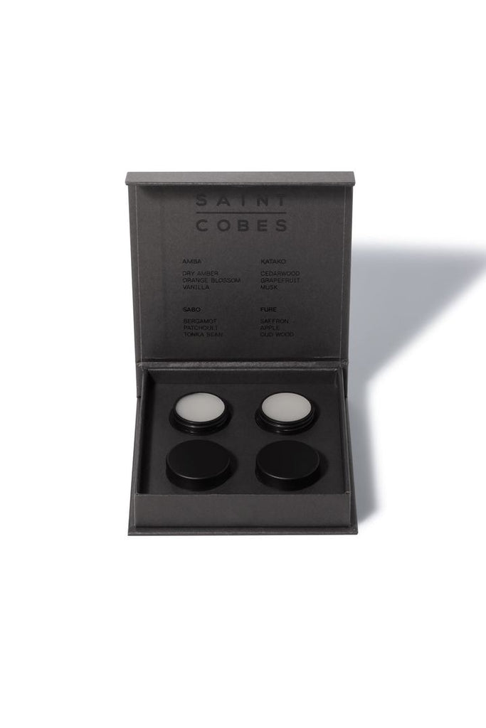 Saint Cobes Discovery Set. Set of four round black pottles containing solid fragrances sitting inside the black Saint Cobes presentation. Two of the pottles have their lids off to show the solid fragrance within.  Presented on a white background