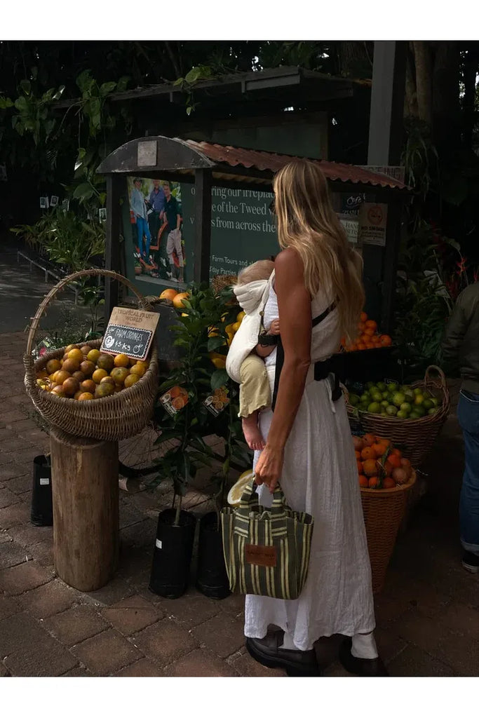 Woman with a child at an outdoor market with fruit stands holding a striped bag