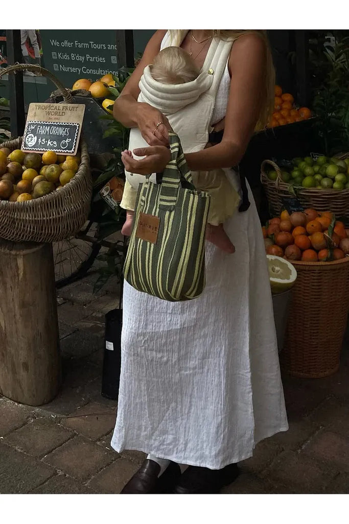 Woman holding a baby in a sling with a striped bag, standing in front of a fruit stand.