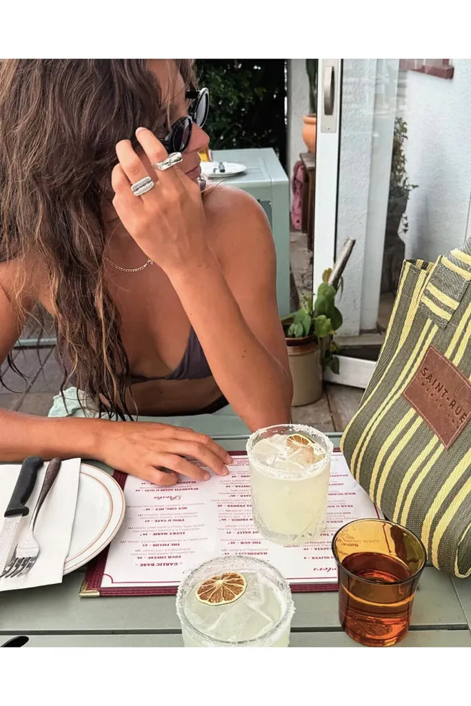 Woman sitting at a table with drinks and a menu, wearing sunglasses and rings with a striped bag beside her