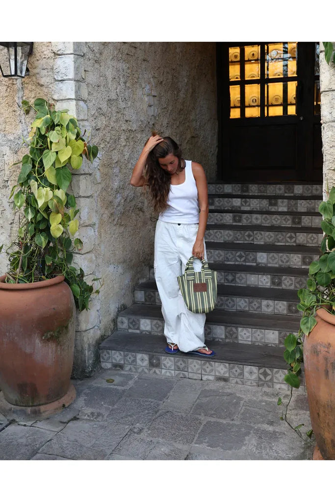 Woman in white outfit standing on stone steps with plants and a door in the background holding a striped bag