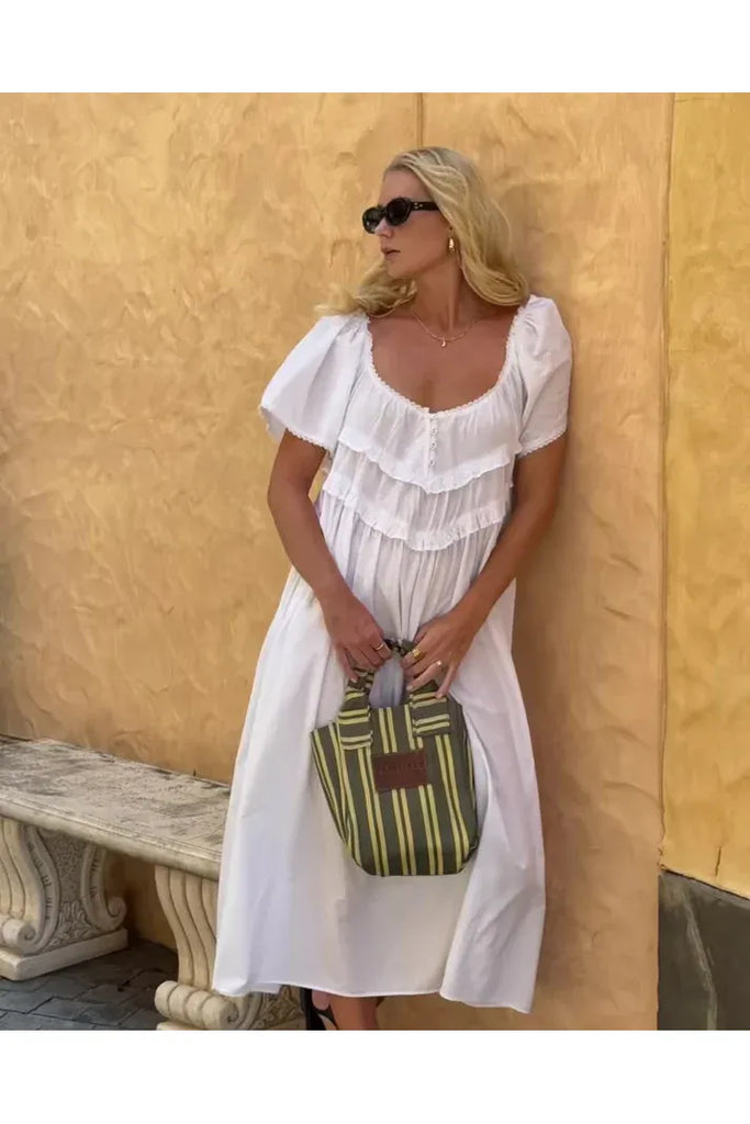 Woman in a white dress holding a striped bag against a yellow wall.