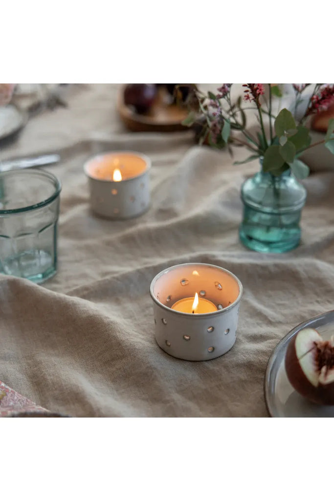 Two lit tealight candles in decorative holders on a textured fabric surface with a vase of flowers and glassware.