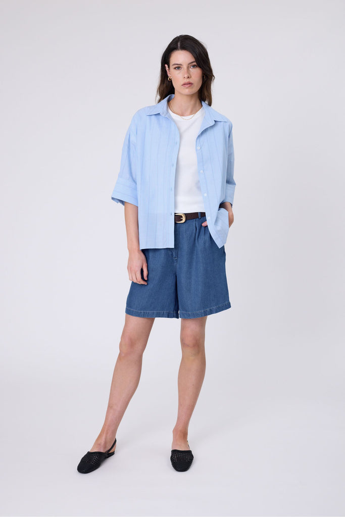 Woman wearing a light blue striped shirt and denim shorts on a white background. Marlow Shoreline Shirt.