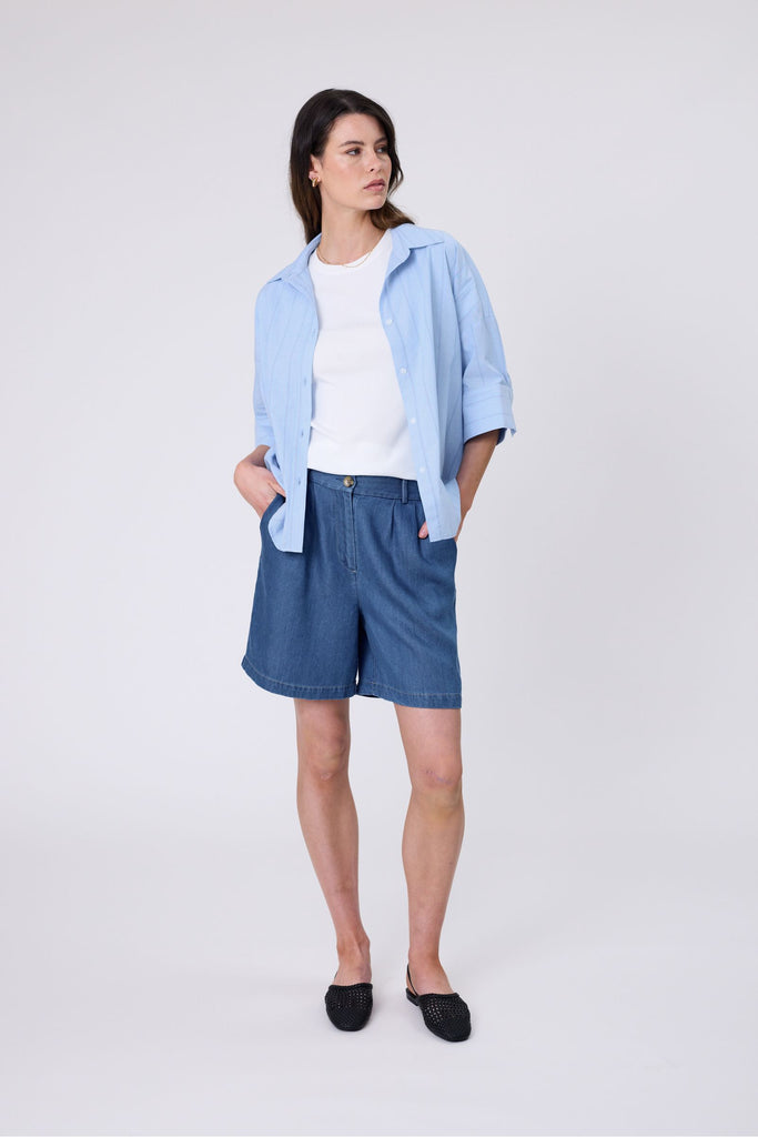 Woman wearing a light blue striped shirt and denim shorts on a white background. Marlow Shoreline Shirt.