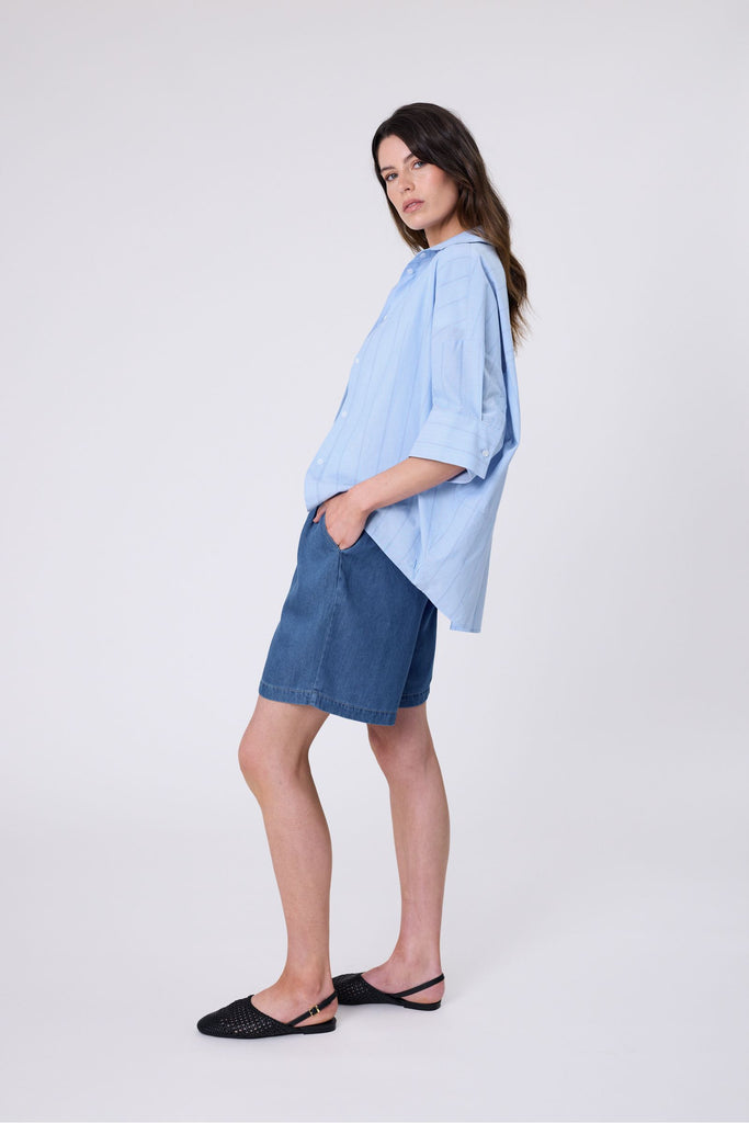 Woman wearing a light blue striped shirt and denim shorts on a white background. Marlow Shoreline Shirt.