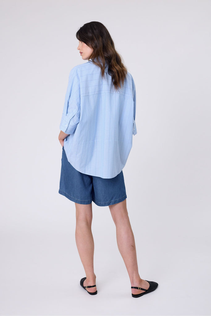 Woman wearing a light blue striped shirt and denim shorts on a white background. Marlow Shoreline Shirt.