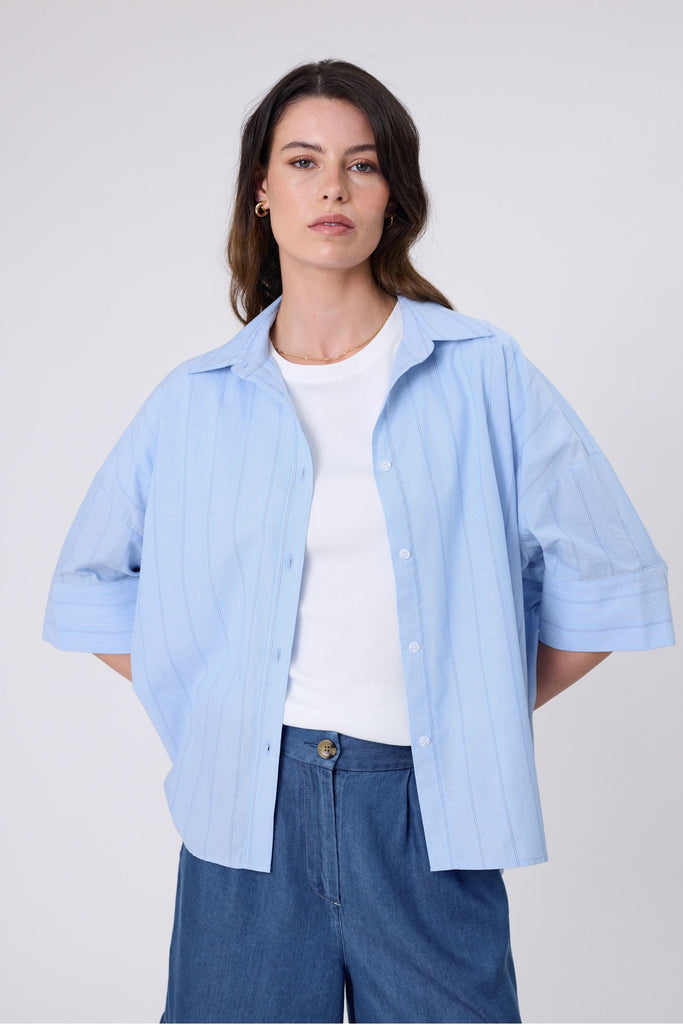 Woman wearing a light blue striped shirt white tee and denim shorts on a white background. Marlow Shoreline Shirt.