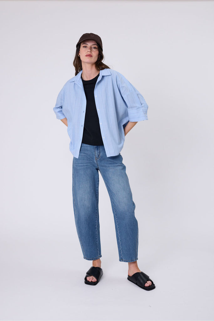 Woman wearing a light blue striped shirt black tee and denim jeans on a white background. Marlow Shoreline Shirt.