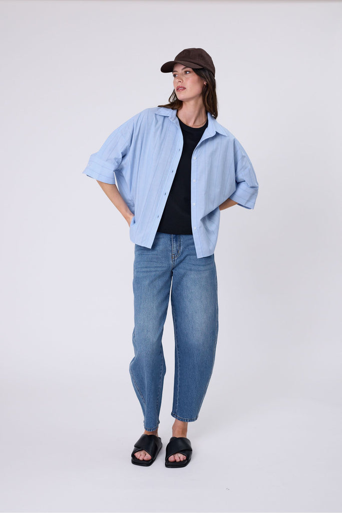 Woman wearing a light blue striped shirt black tee and denim jeans on a white background. Marlow Shoreline Shirt.