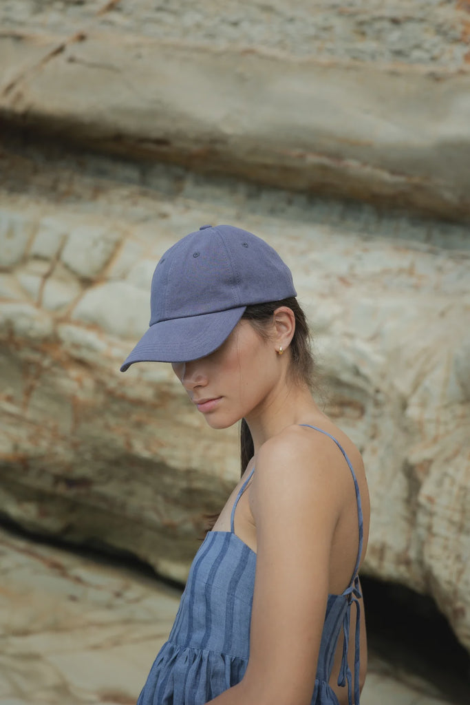 Woman wearing a blue cap and dress standing against a rocky background. Sophie Twill Cap in Denim.