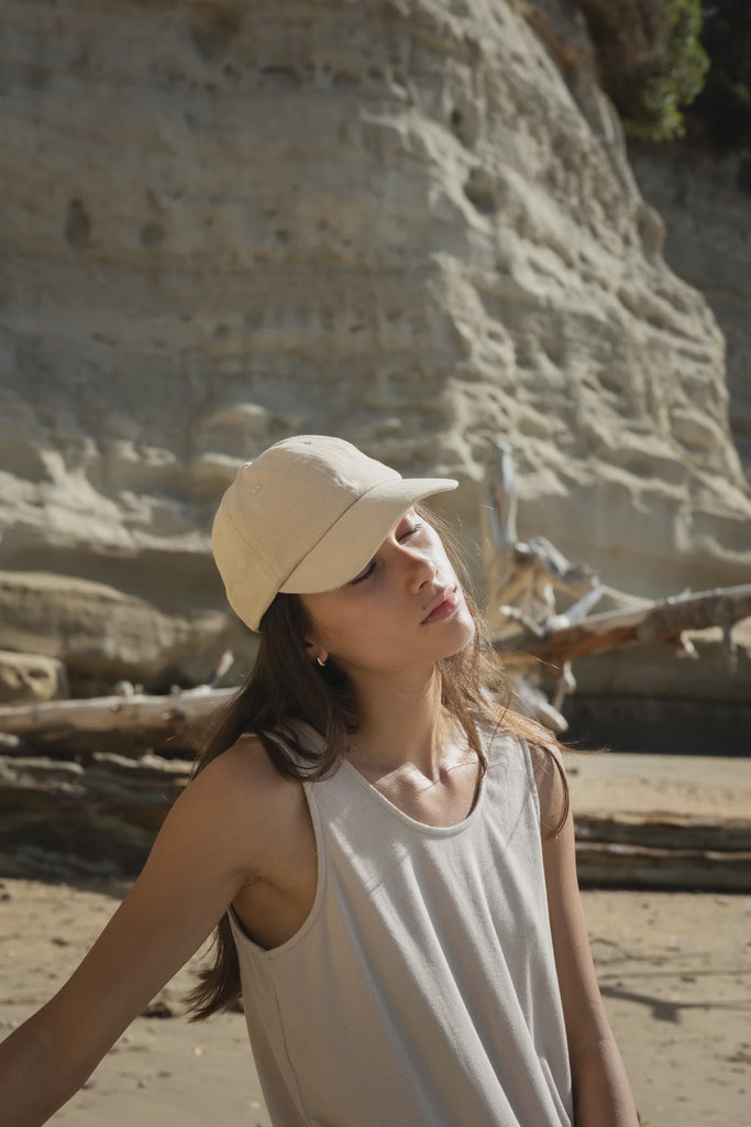 Woman wearing a beige cap and white tank top standing on a beach with cliffs in the background. Sophie Twill cap in stone.