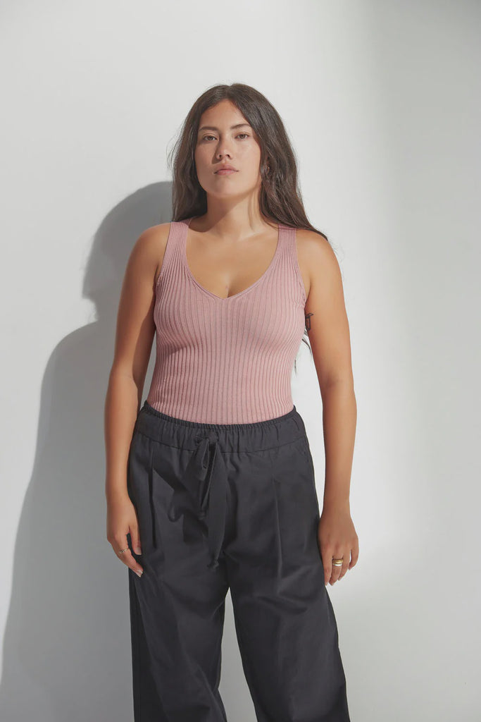 Woman wearing a pink tank top and dark pants against a plain background. Standard Issue Merino Rib V Neck Tank in Coral.