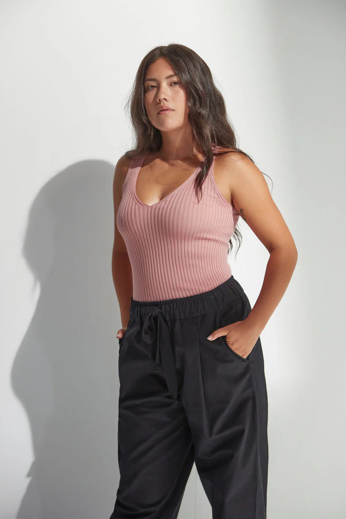Woman wearing a pink tank top and dark pants against a plain background. Standard Issue Merino Rib V Neck Tank in Coral.