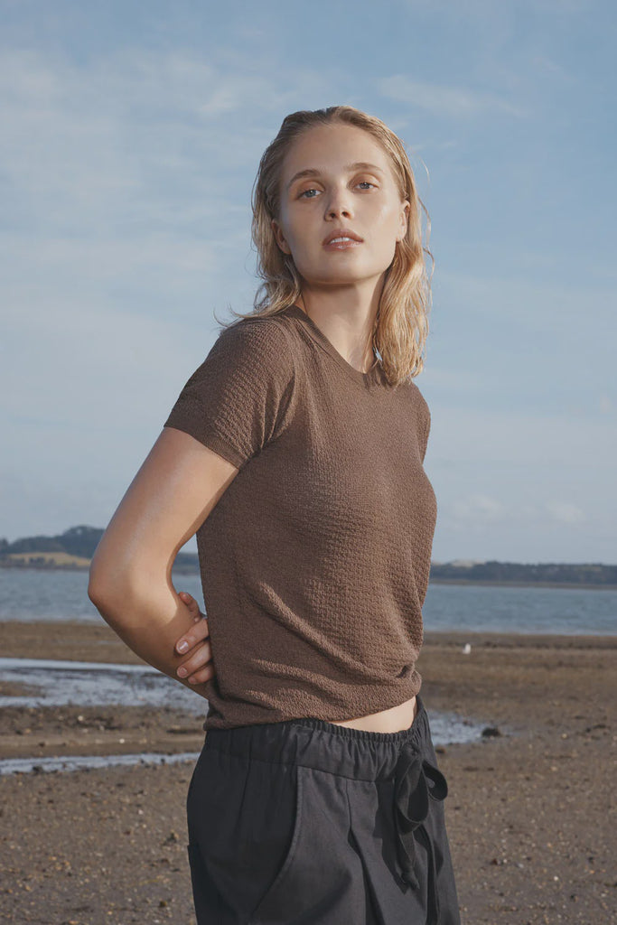 Woman standing on a beach wearing a brown t-shirt and dark pants with a blue sky and water in the background. Standard Issue Seta Picot Tee in Anchor brown.