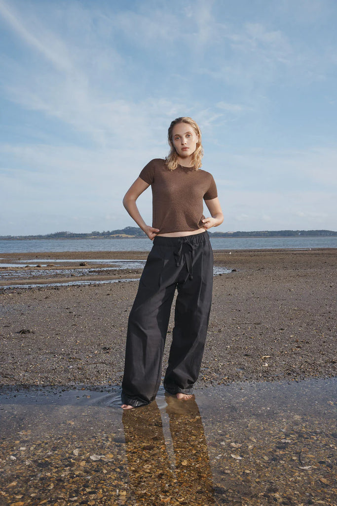 Woman standing on a beach wearing a brown t-shirt and dark pants with a blue sky and water in the background. Standard Issue Seta Picot Tee in Anchor brown.
