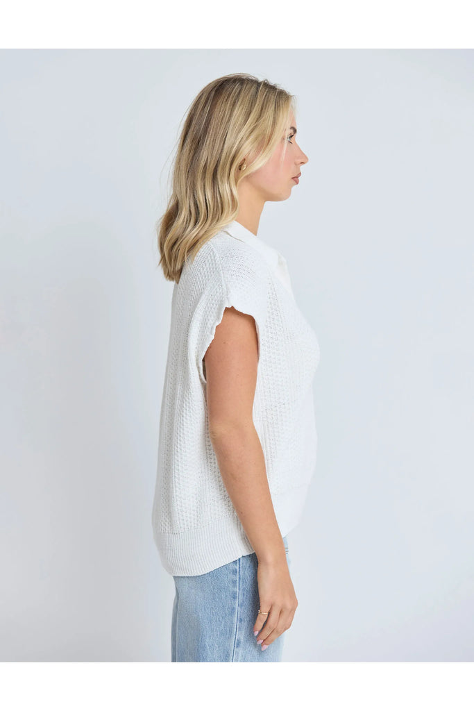 Woman wearing a white sleeveless sweater and light blue jeans against a plain background.