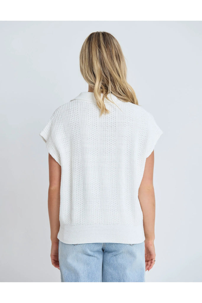 Woman wearing a white sleeveless sweater and light blue jeans against a plain background.