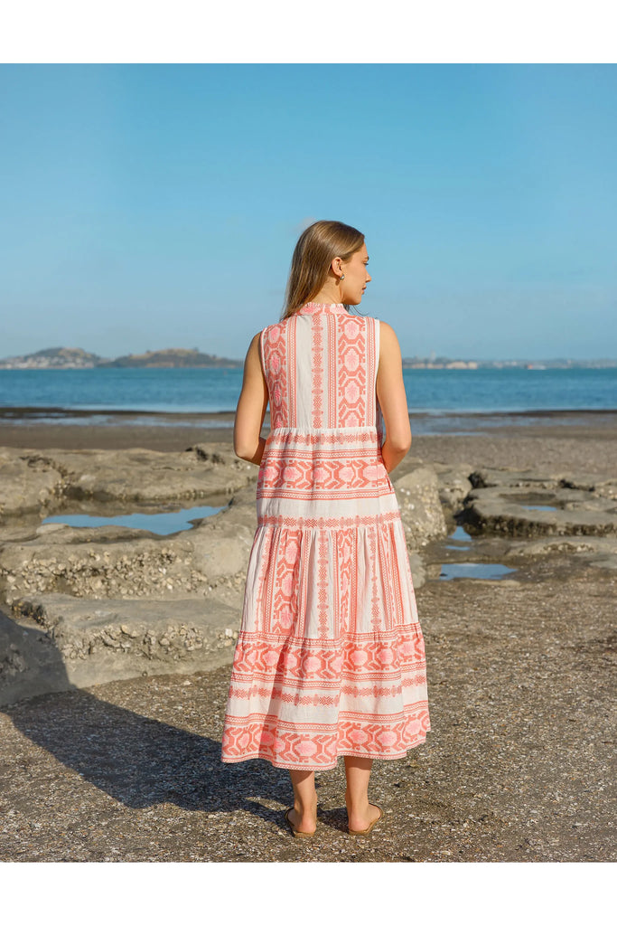Woman in a pink and white patterned dress standing on a beach with ocean and sky in the background