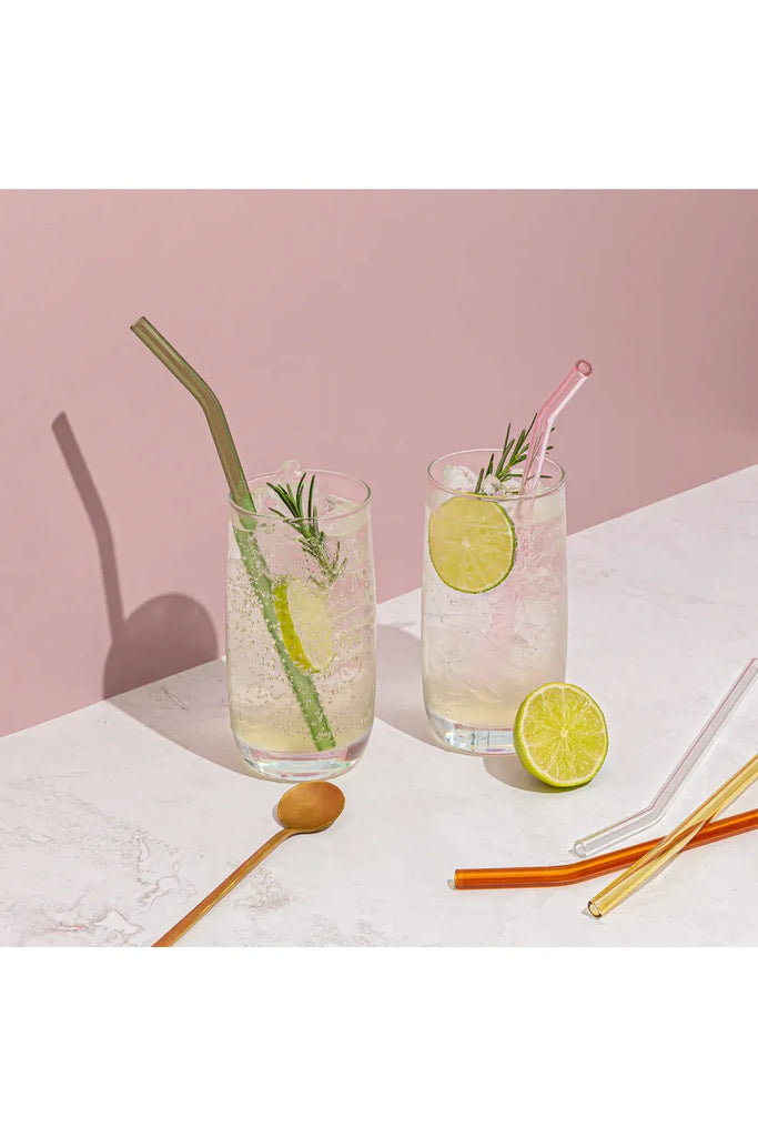 Two glasses of lemonade with lime slices and rosemary, and a Sucker Reusable glass straw on a pink background.