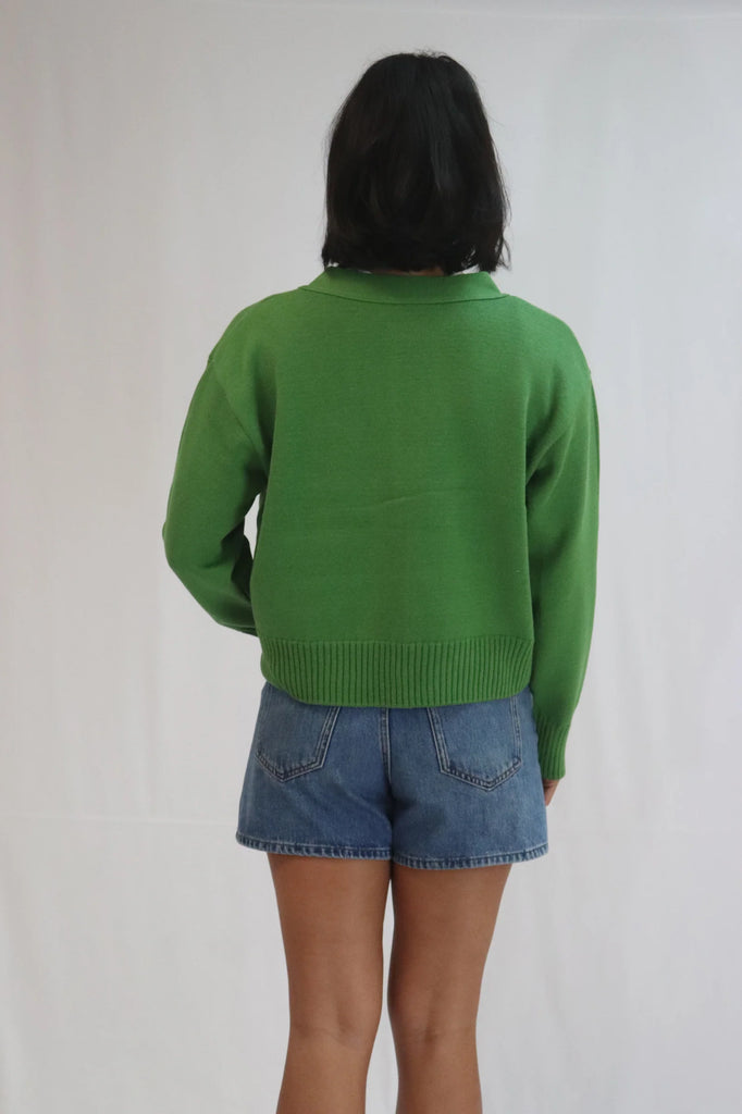 Person wearing a green cardigan over a white shirt with blue jeans on a plain background. Tatano Solid Cardi in herb green.
