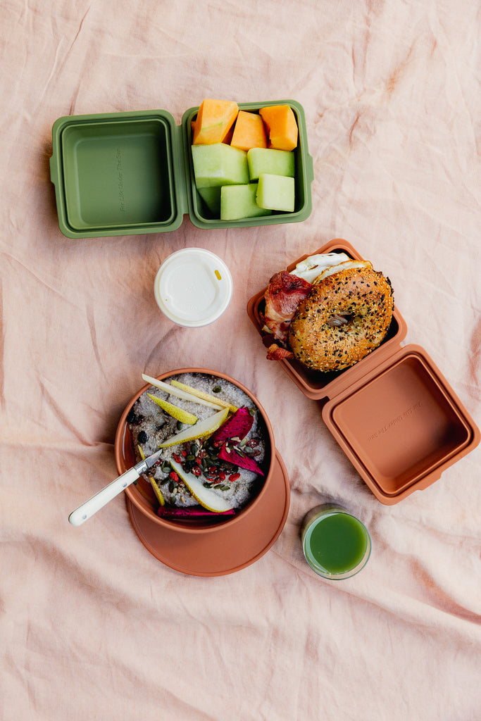 TOGO Sun The Burger Box in Spice. Displayed as part of a breakfast on the run image.  Image shows The Burger Box in both the the Spice and Cactus open and filled with melon and a Bagel.  Sitting alongside is the TOGO Sun Bowl with fruit and granola along with a takeaway coffee and green juice