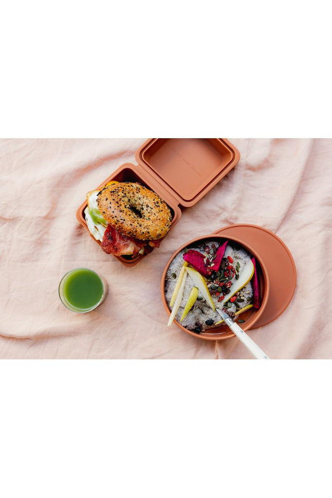 TOGO Sun The Burger Box in Spice. Displayed open and filled with a bacon & egg bagel sitting alongside the Togo Bowl in Spice filled with fruit and granola and a glass of green juice.