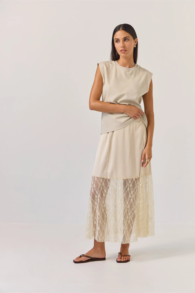 Woman wearing a beige sleeveless tank top and lace skirt on a plain background. Tuesday Noon Tank in Cement.