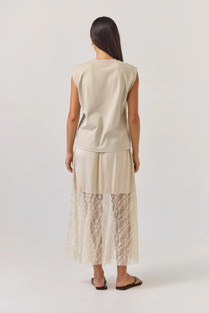 Woman wearing a beige sleeveless tank top and lace skirt on a plain background. Tuesday Noon Tank in Cement.