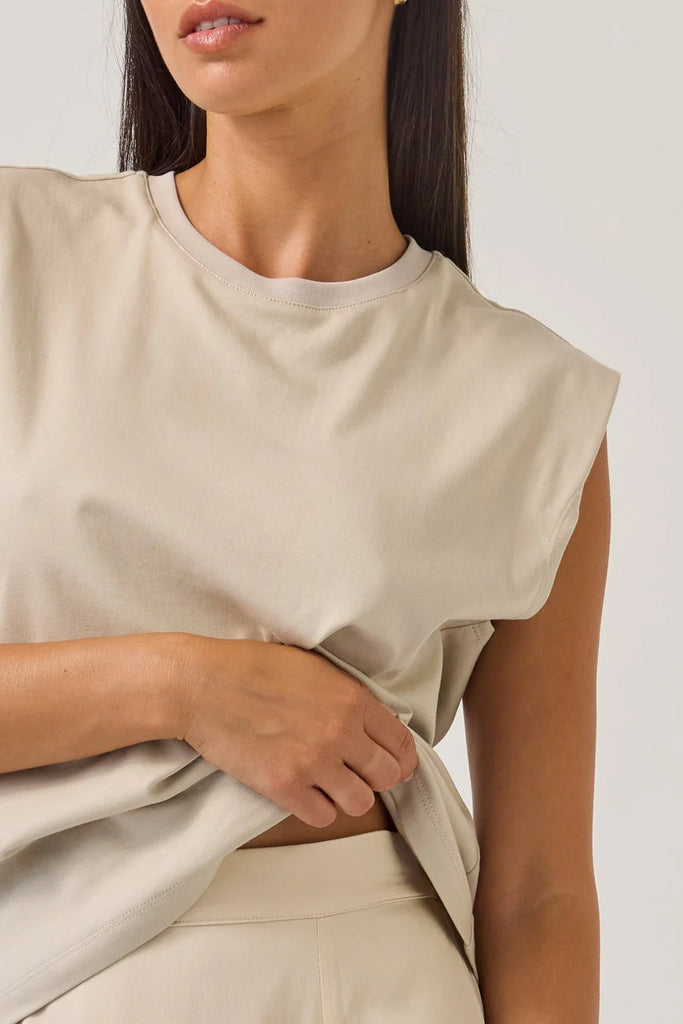 Woman wearing a beige sleeveless tank top on a plain background. Tuesday Noon Tank in Cement.
