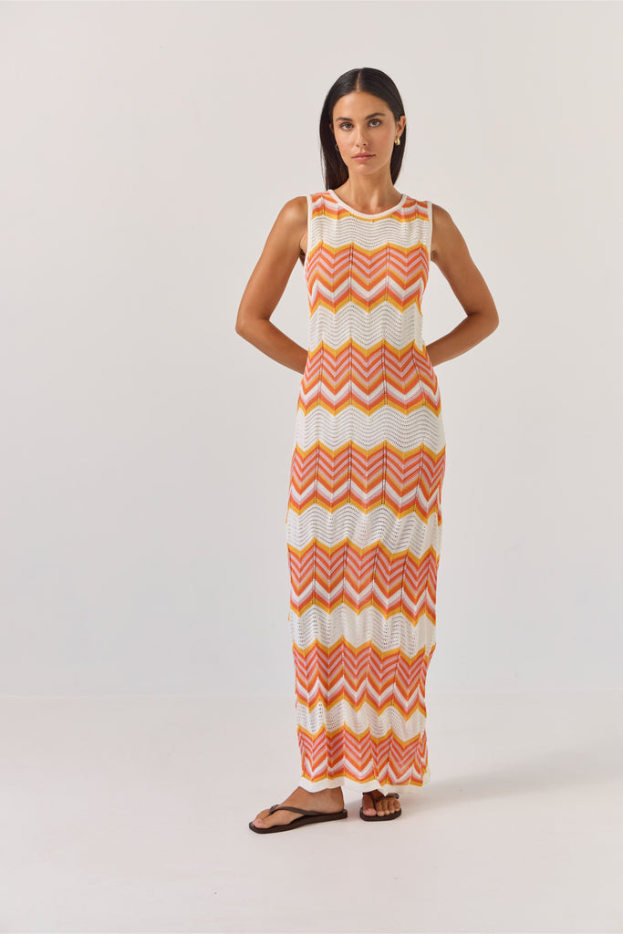 Woman wearing a sleeveless dress with orange and white zigzag pattern on a plain background. Tuesday Label Horizon Dress in a chevron pattern.