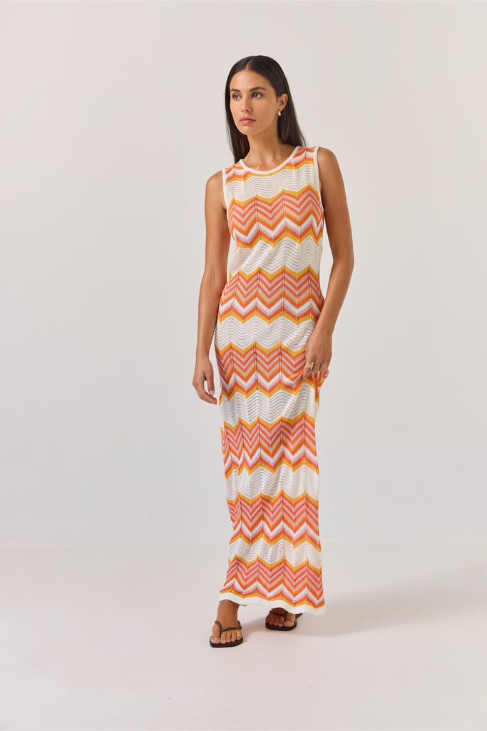 Woman wearing a sleeveless dress with orange and white zigzag pattern on a plain background. Tuesday Label Horizon Dress in a chevron pattern.