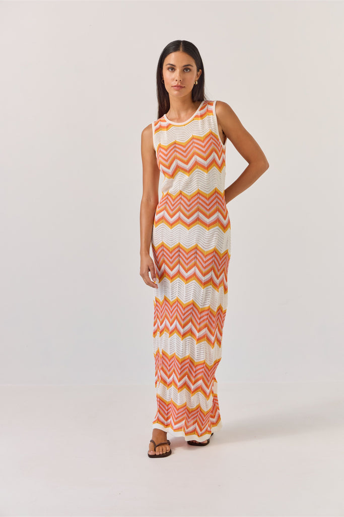 Woman wearing a sleeveless dress with orange and white zigzag pattern on a plain background. Tuesday Label Horizon Dress in a chevron pattern.