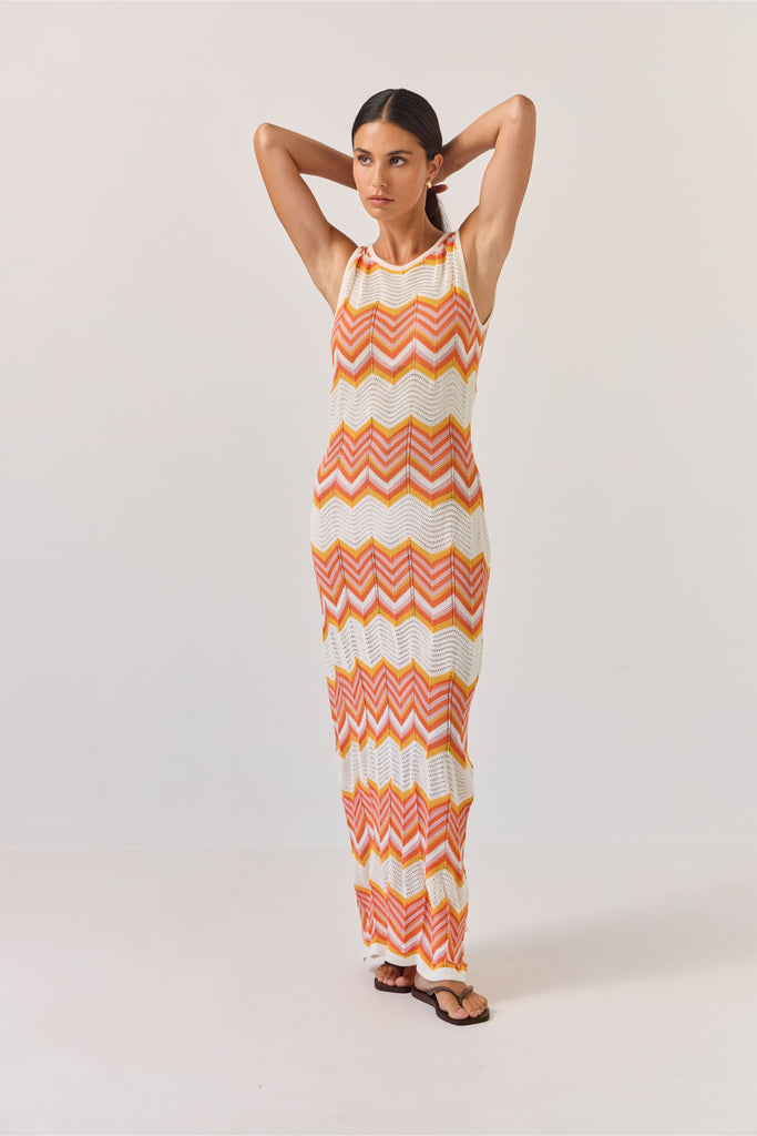 Woman wearing a sleeveless dress with orange and white zigzag pattern on a plain background. Tuesday Label Horizon Dress in a chevron pattern.