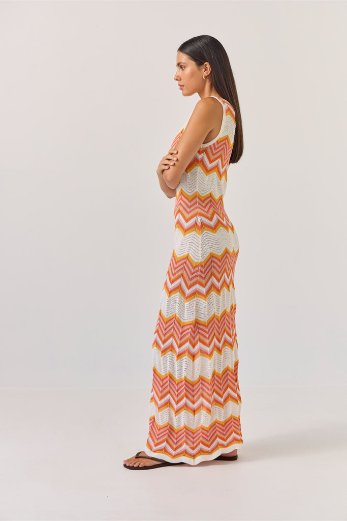 Woman wearing a sleeveless dress with orange and white zigzag pattern on a plain background. Tuesday Label Horizon Dress in a chevron pattern.