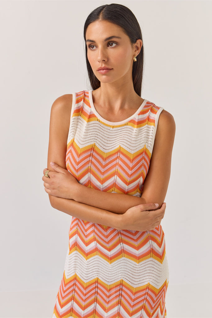 Woman wearing a sleeveless dress with orange and white zigzag pattern on a plain background. Tuesday Label Horizon Dress in a chevron pattern.