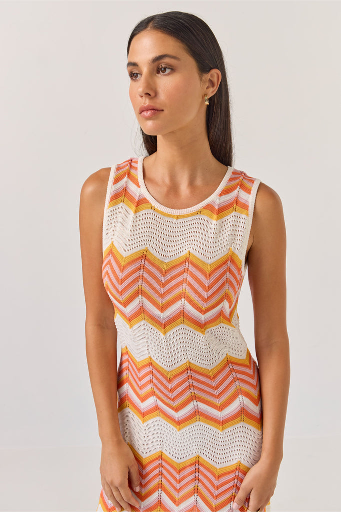 Woman wearing a sleeveless dress with orange and white zigzag pattern on a plain background. Tuesday Label Horizon Dress in a chevron pattern.