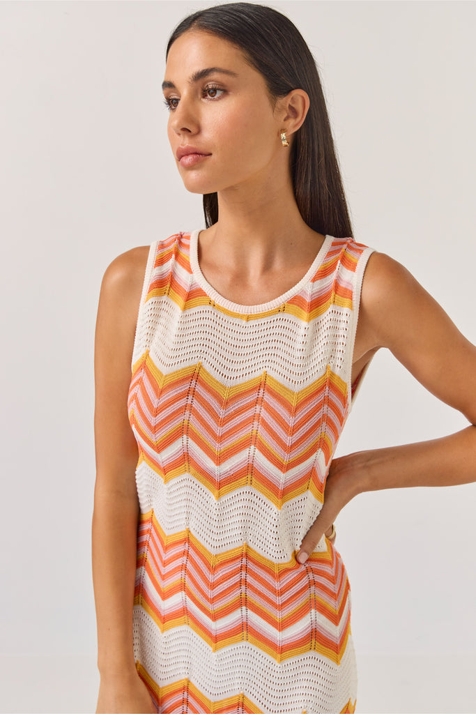 Woman wearing a sleeveless dress with orange and white zigzag pattern on a plain background. Tuesday Label Horizon Dress in a chevron pattern.