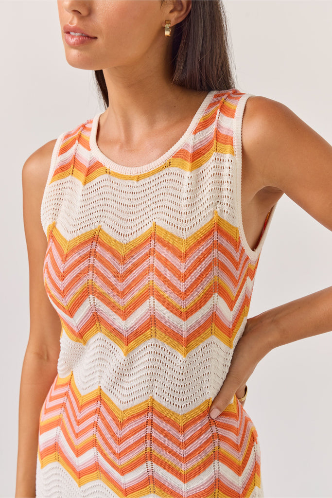 Woman wearing a sleeveless dress with orange and white zigzag pattern on a plain background. Tuesday Label Horizon Dress in a chevron pattern.