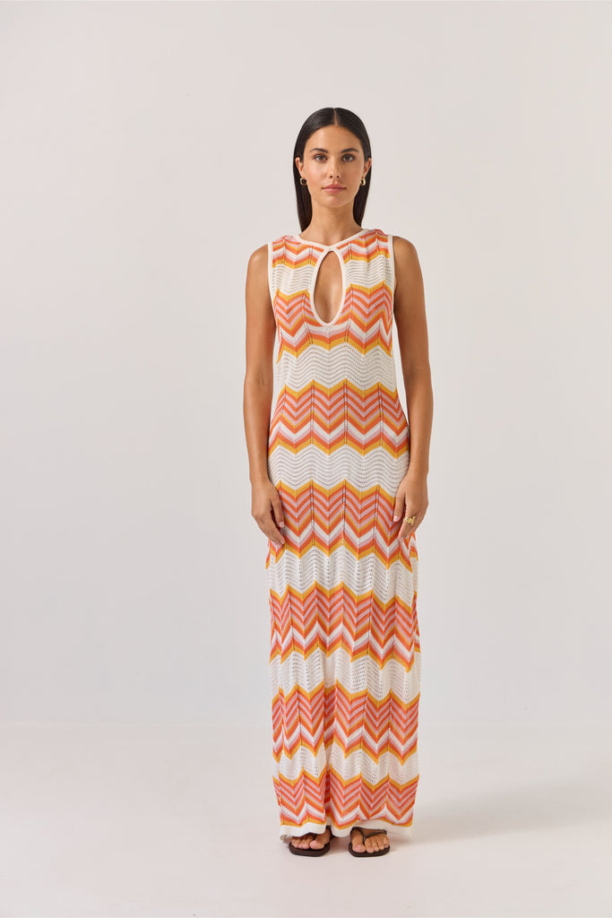 Woman wearing a sleeveless dress with orange and white zigzag pattern on a plain background. Tuesday Label Horizon Dress in a chevron pattern.