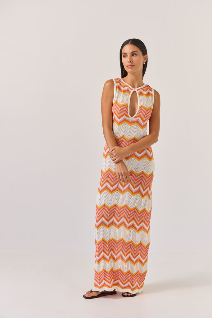 Woman wearing a sleeveless dress with orange and white zigzag pattern on a plain background. Tuesday Label Horizon Dress in a chevron pattern.
