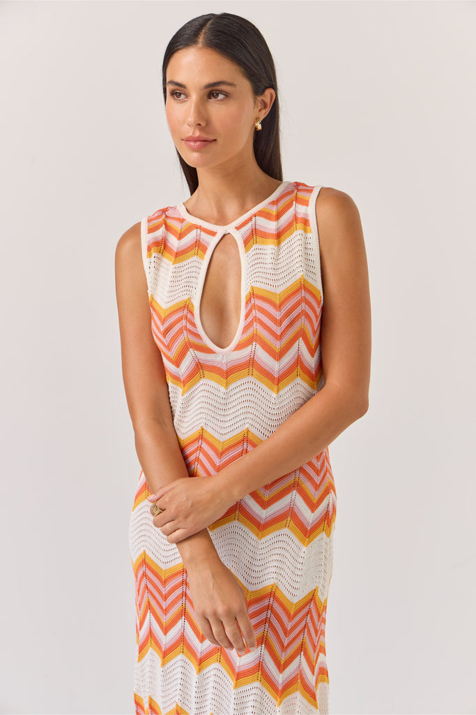 Woman wearing a sleeveless dress with orange and white zigzag pattern on a plain background. Tuesday Label Horizon Dress in a chevron pattern.